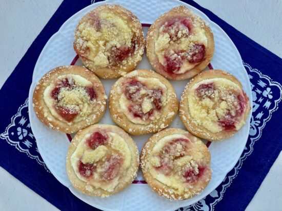 Talíř s kynutými koláči s tvarohovou náplní, jahodami a drobenkou, položený na bílo-modrém ubrusu s lidovým vzorem.